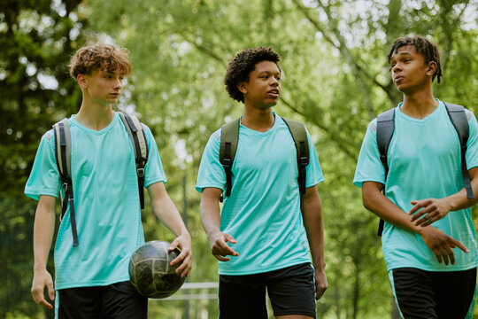 Three teenage boys, two Black and one Caucasian, walking outdoors with backpacks and soccer ball, engaging in conversation after sports practice in green park setting - Powered by Adobe