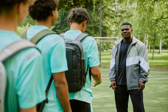 Black man coach standing on sports field talking to group of teenage boys with backpacks, teenagers listening attentively outdoors, trees and soccer goal visible in background - Powered by Adobe