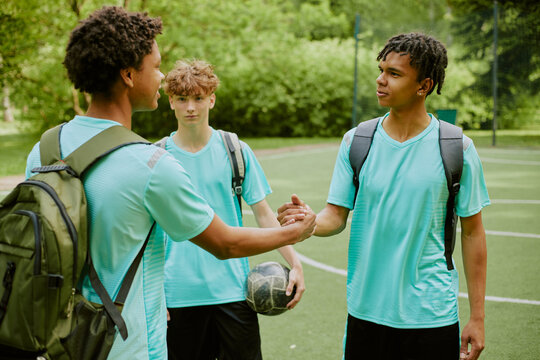 Two Black teenage boys shaking hands while standing on outdoor sports field, third Caucasian teenage boy holding soccer ball and watching interaction, all wearing athletic shirts and backpacks