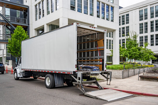 Day cab middle duty white semi truck with open box trailer and lowered lift standing on the urban city street with multilevel apartments unloading delivered goods