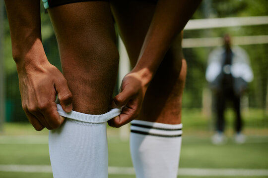 Person adjusting soccer socks on field, hands pulling up athletic socks with goalpost and blurred figure in background, focusing on preparation before sports activity