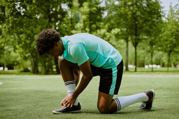Black teenager tying shoelaces on sports field, preparing for athletic activity outdoors, wearing...