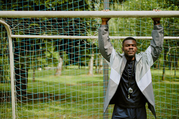 Black young adult man hanging from soccer goalpost with both hands outdoors, wearing sports jacket...