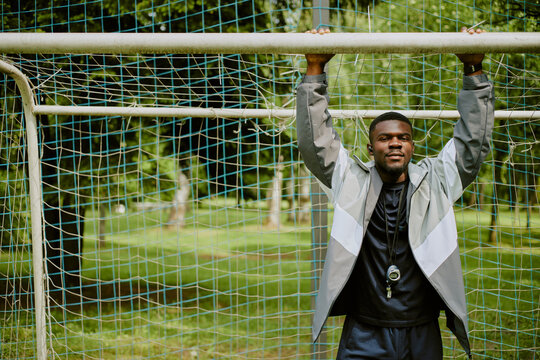 Black young adult man hanging from soccer goalpost with both hands outdoors, wearing sports jacket and whistle around neck, standing on grassy field with trees in background - Powered by Adobe