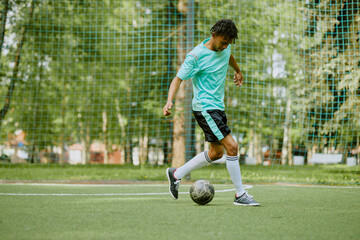 Teenage boy dribbling soccer ball on outdoor field, focusing on footwork during practice session, netted goal and green trees visible in background, athletic movement captured