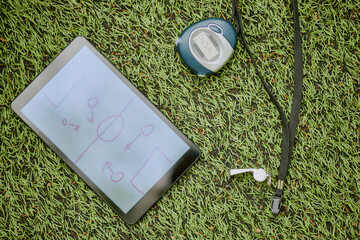 Tablet displaying soccer field tactics diagram lying on artificial grass next to digital stopwatch and black whistle with lanyard, representing sports coaching equipment