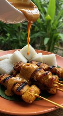 Chicken satay skewers on a plate with rice cakes, being drizzled with peanut sauce, against a green leafy background.
