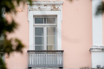 Ornate balcony with a vintage window on a pastel pink building facade, framed by blurred greenery