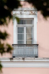 Ornate balcony with a vintage window on a pastel pink building facade, framed by blurred greenery