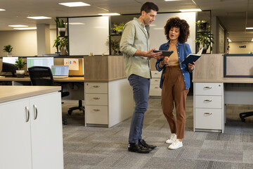 Reviewing data, Diverse colleagues standing by desks at open-plan office with tablet and notebook