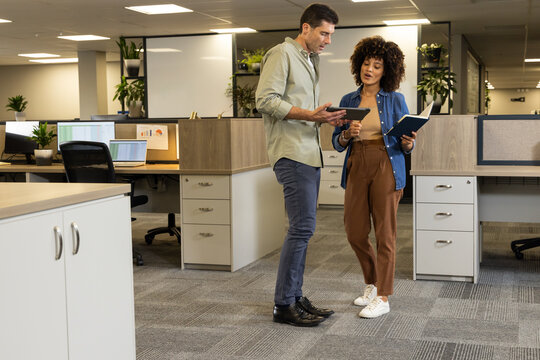 Reviewing data, Diverse colleagues standing by desks at open-plan office with tablet and notebook