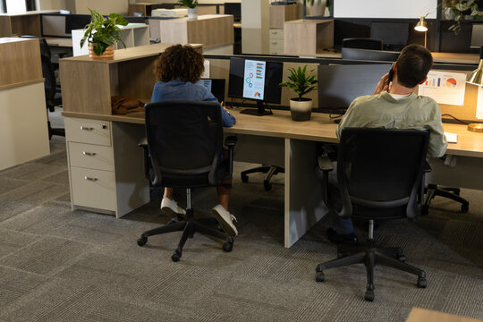 Working Diverse coworkers typing at wooden desks in open-plan office, with dual monitors - Powered by Adobe