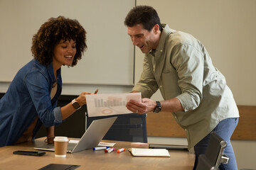 Analyzing Diverse coworkers studying bar chart at office table, with laptop smartphone coffee cup