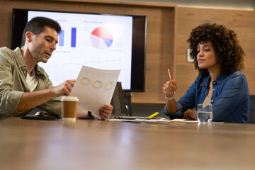 Reviewing Diverse coworkers analyzing printed report at office conference room, with monitor graphs
