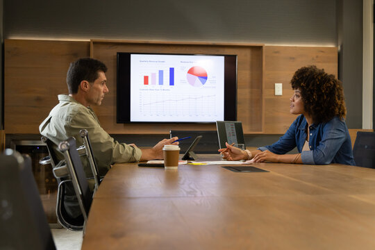 Diverse coworkers collaborating at wall-mounted display in conference room, with laptops and pens