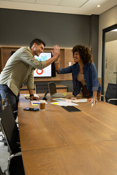 Diverse coworkers high-fiving at glass-walled conference room, with laptops and printed charts