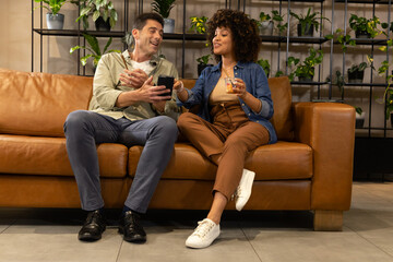 Chatting diverse coworkers sitting on brown leather sofa in lounge, with smartphone and glass cup