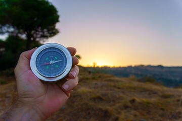Hand is holding a compass showing direction at sunset in the nature