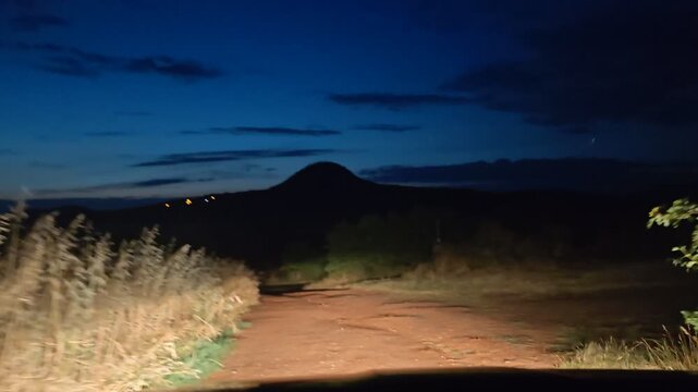 Night offroad drive on muddy bumpy country road with passenger car and mountain silhouette in the background POV of driver with lights shining through the dark night