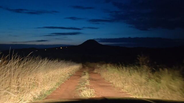 Night offroad drive on muddy bumpy country road with passenger car and mountain silhouette in the background POV of driver with lights shining through the dark night