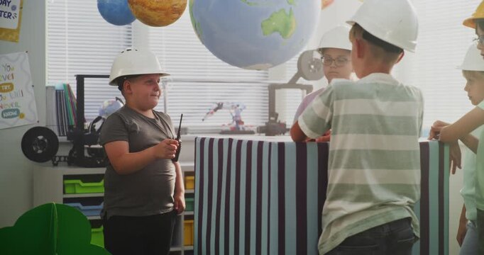 Team of Primary School Students in Hardhats Discussing City Planning, Young Boy Giving Instructions. Interactive STEM Class with Large Cardboard Urban Blocks and Models of Trees, Buildings. Dolly Shot
