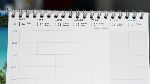 Close-up of a Hungarian weekly calendar showing the days from Monday to Saturday, with names associated with each date. A person is flipping the page, revealing part of the next week.