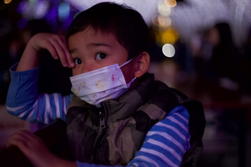A young boy wearing a striped shirt, puffy vest, and a cute printed face mask looks curiously at...