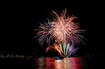 Vibrant fireworks explode over a calm water body, reflecting colorful lights in the night sky.