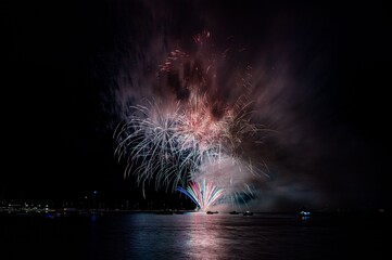 Vibrant fireworks explode over a calm water body, reflecting colorful lights in the night sky.