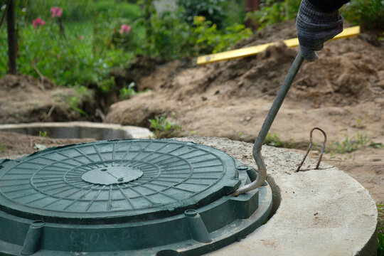 Opening a green septic tank manhole cover with a metal hook in a private house yard for inspection and maintenance of a local sewage system, wastewater management, and underground drainage infrastruct