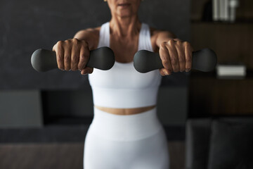 Middle aged Caucasian woman exercising with dumbbells, extending both arms forward while standing...