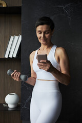 Middle aged Caucasian woman standing holding dumbbell in one hand while using smartphone with other...