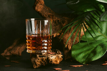 Blended whiskey in glass. Dark background with old driftwood and big leaves