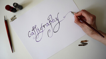 Calligraphy process. Female hand writing word Calligraphy. Handwritten font. Pens, ink are lying...