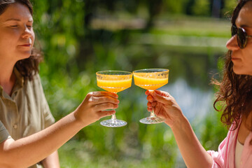 Two women seating on a picnic blanket by a lakeside, toasting with orange beverages in glasses. A...