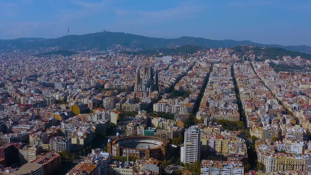 4K drone panorama of Barcelona Eixample: precise grid converging on the Sagrada Familia, La Monumental bullring below, dense rooftops, tree lined avenues, and Collserola hills under clear blue sky.