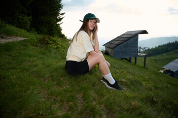Woman hiker sitting on the hill in the mountains early in the morning near wooden cabins. Peaceful morning in nature, thoughtful expression, natural beauty and golden light.