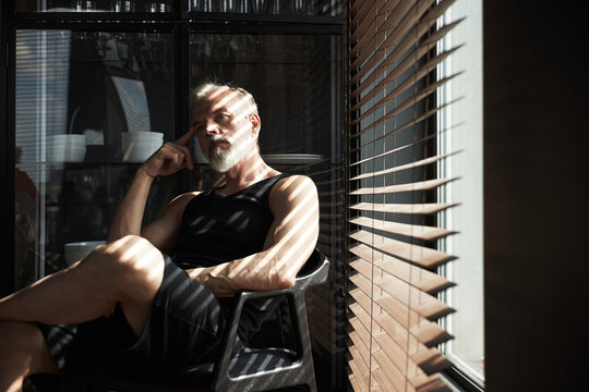 Caucasian senior man sitting in chair resting head on hand gazing thoughtfully, window with sunlight casting striped shadows across face and body through blinds