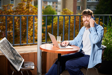Man works on laptop and smartphone at outdoor table, with glass of iced coffee. Solar panel nearby, emphasizing sustainable, modern eco-friendly workspace that combines technology and sustainability.