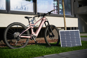 Pink electric mountain bike parked on grassy lawn, connected to solar panel for charging. Bike features rugged tires and positioned beside building, with its shadow cast on wall.