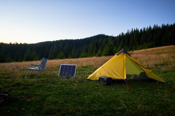Tourist tent with photovoltaic solar panel and portable power station nearby in grassy field at sunset. Scene surrounded by rolling hills under clear blue sky, showcasing eco-friendly camping setup.