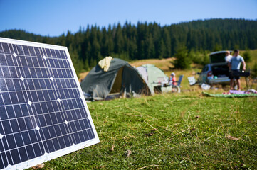 Close-up solar panel in grassy field with tents in background highlights eco-friendly camping setup. Scene includes campers and car, emphasizing integration of renewable energy in outdoor activities.