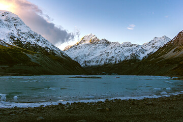 Icy alpine lake beneath towering snowy peaks, glowing in soft sunset light.