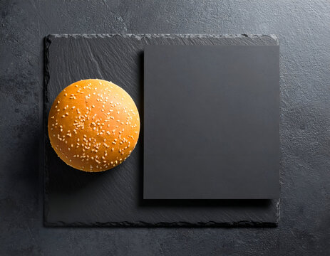 Overhead shot of a golden sesame seed burger bun resting on a dark black slate plate next to a versatile blank mockup card, perfect for showcasing your culinary designs or branding. - Powered by Adobe