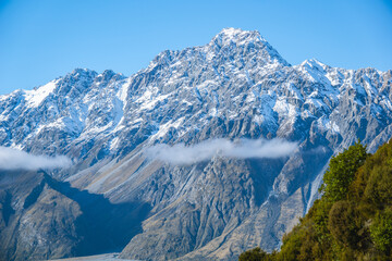 Rugged snow-covered mountain rises above a valley, partially veiled by thin clouds.