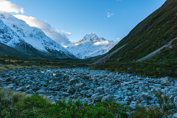 Towering snowy peak rises above a rocky valley surrounded by green and alpine slopes.