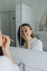 Middle-aged woman applying eye makeup with a brush in front of a bathroom mirror. Beauty routine...