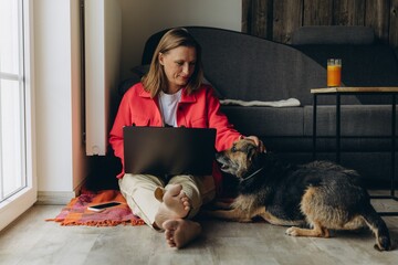 Freelancer woman working from home with laptop, sitting on floor in cozy living room, barefoot and...