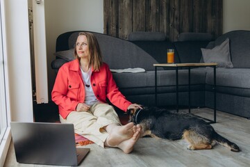 Freelancer woman working from home with laptop, sitting on floor in cozy living room, barefoot and...