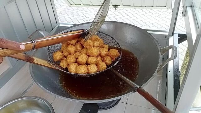 Street food vendor tahu crispy or frying tofu cubes in hot oil, scooping the golden brown pieces with a strainer from a large wok.